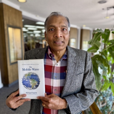 Board member holding a copy of the book The Mobile Wave inside the Library, standing near a railing with plants and bookshelves in the background.