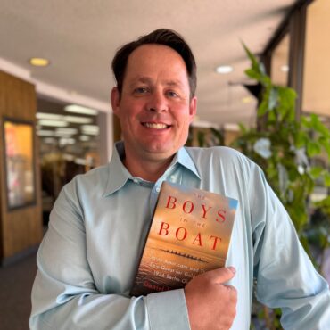 Board member holding a copy of the book "The Boys in the Boat" inside the Library, standing near a railing with plants and bookshelves in the background.