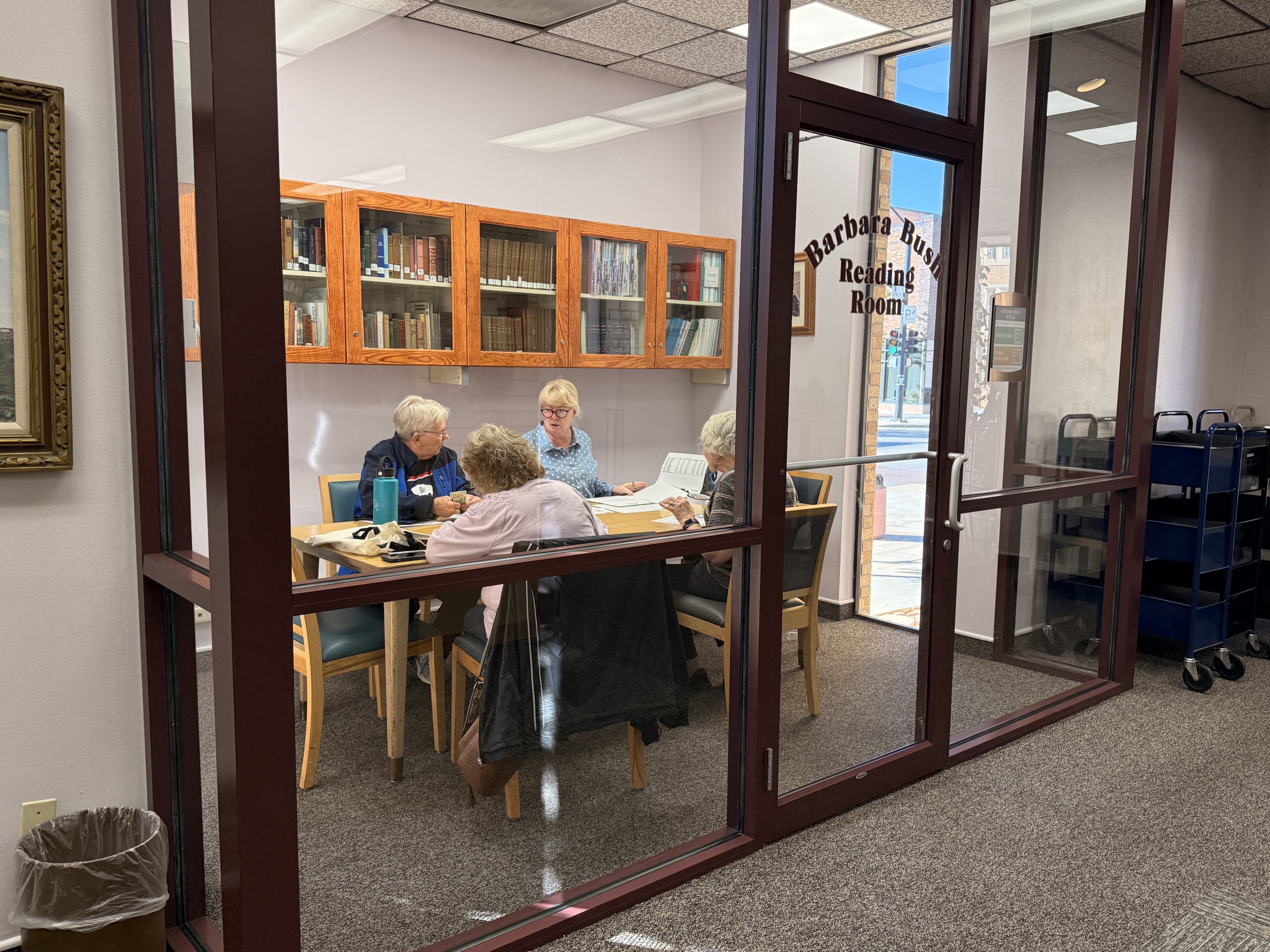 Older adults sit around a table in a glass-walled room at the library, talking and looking at papers together. The door reads “Barbara Bush Reading Room,” and bookshelves line the wall behind them.