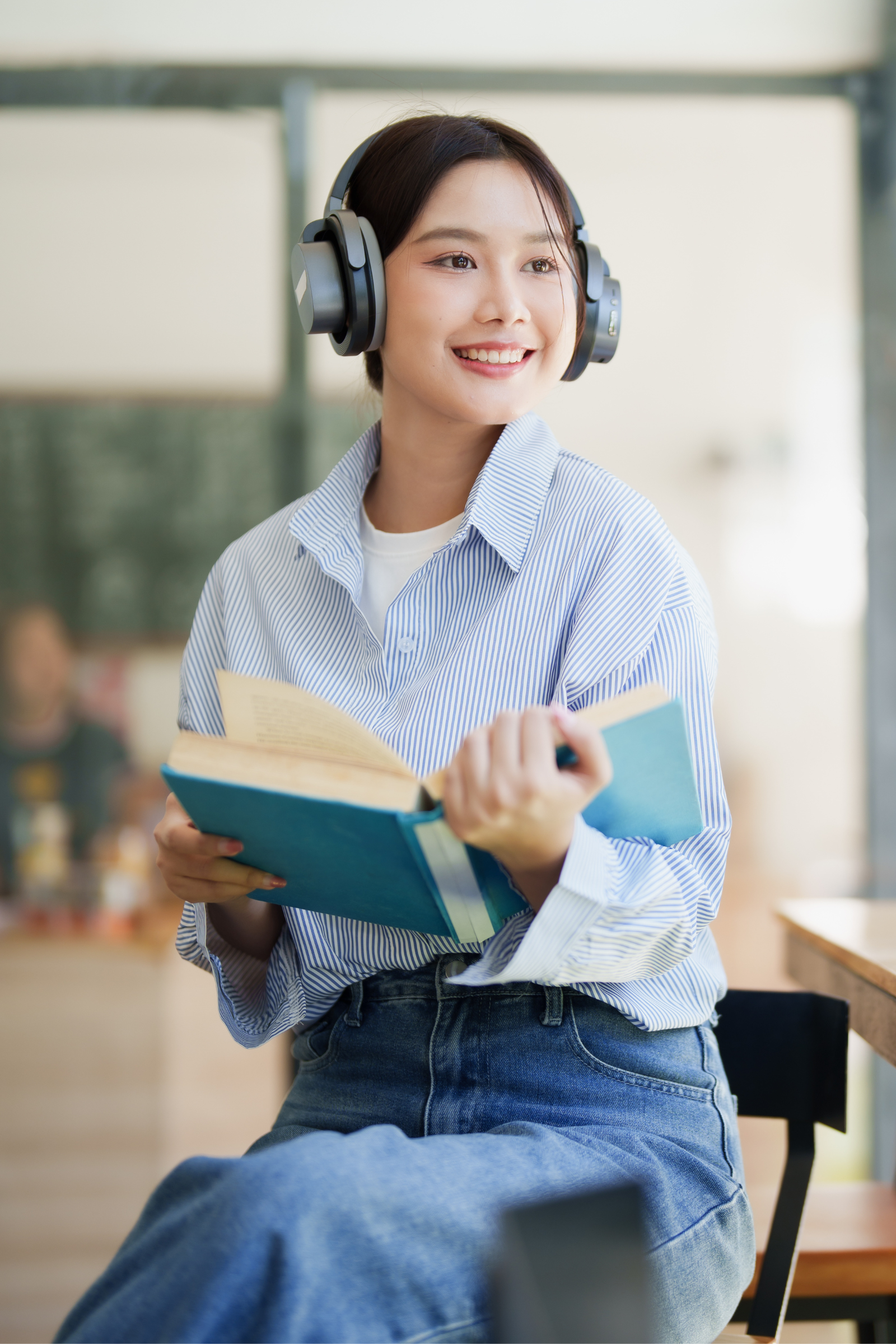 Person wearing over-ear headphones sits at a table in a bright café or library setting, smiling while holding and reading an open book, with natural light and blurred people in the background.