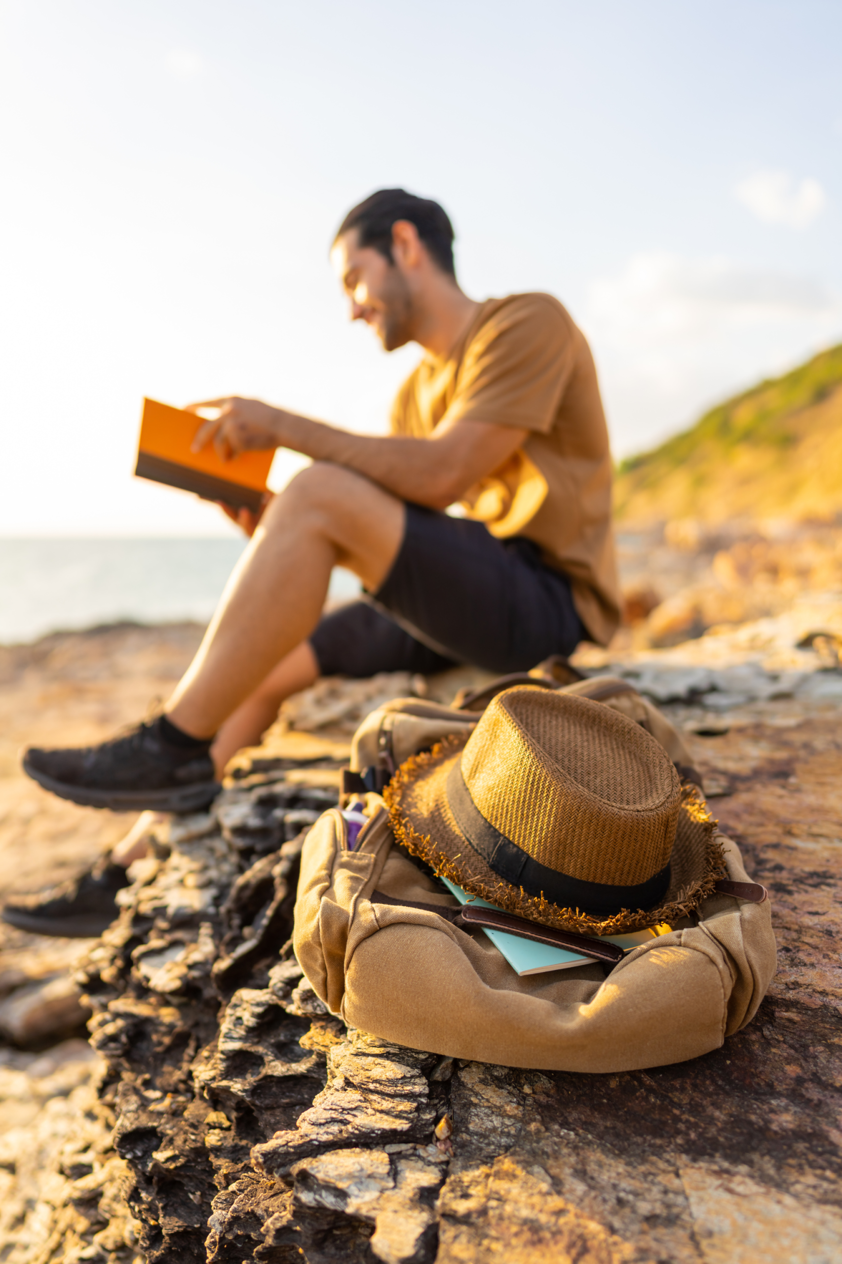 Person sitting on rocky shoreline reading a book at sunset, with a backpack and straw hat resting nearby.