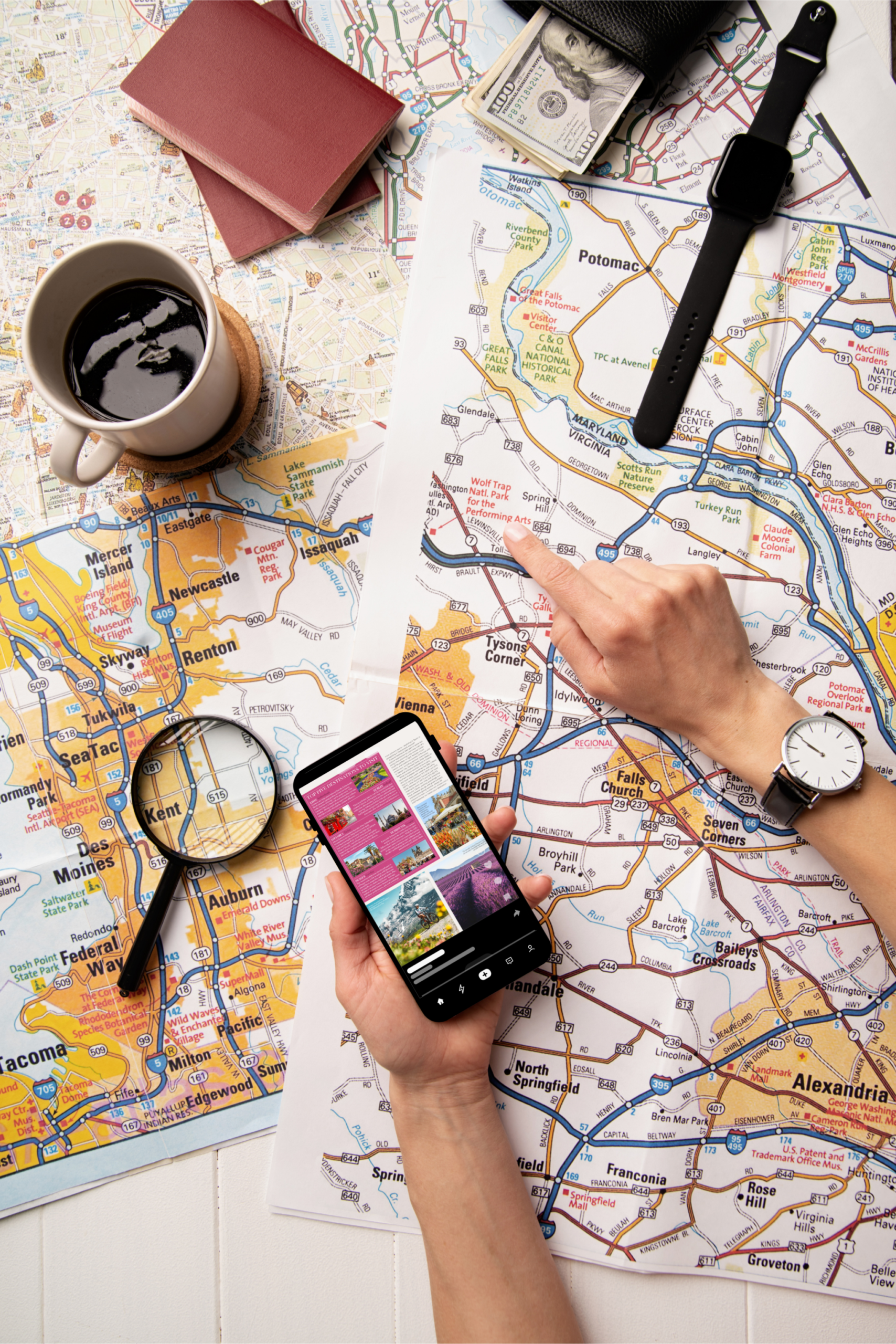 Hands using paper maps and a smartphone to plan a trip, surrounded by passports, cash, a watch, and a cup of coffee.